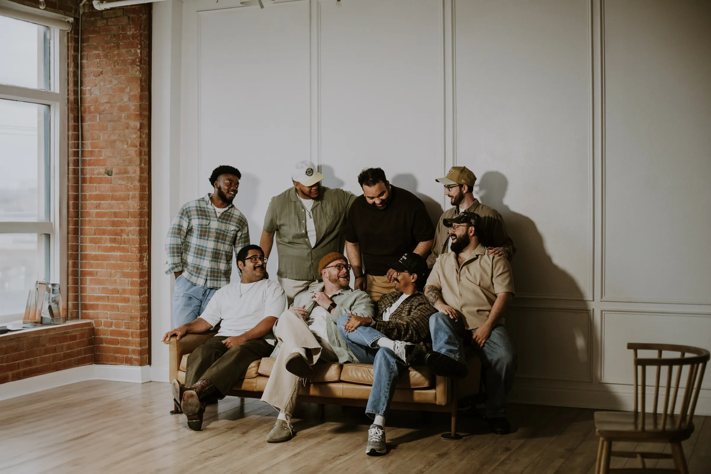 Eight men on a couch under the brick windows, one empty chair