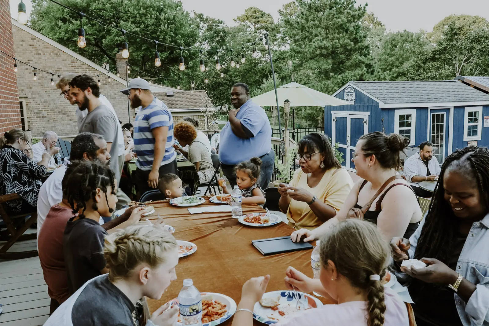 Families sharing dinner at a long table on a patio under string lights
